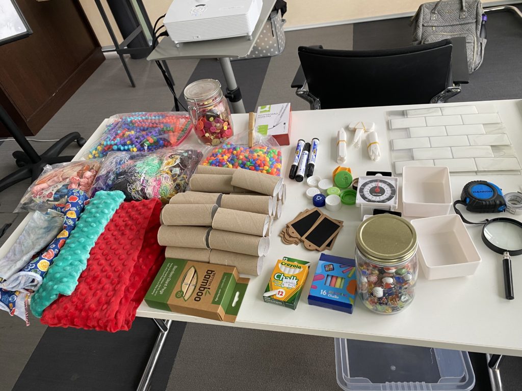 Various crafting items, including toilet tubes, chalk, beads, markers, and buttons are laid out on a table.